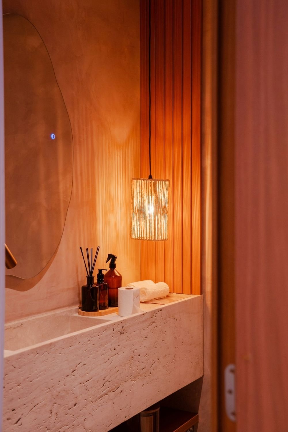 Travertine trough sink with a woven rope pendant light, amber bottles and a wood slat wall in warm light