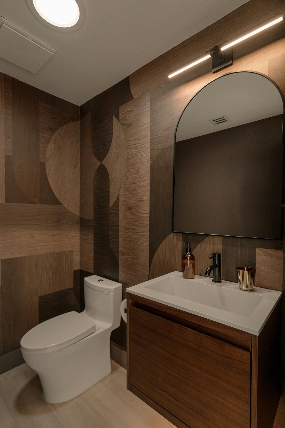 Powder room with wood-look geometric wallpaper, arched black mirror, linear light bar and walnut floating vanity