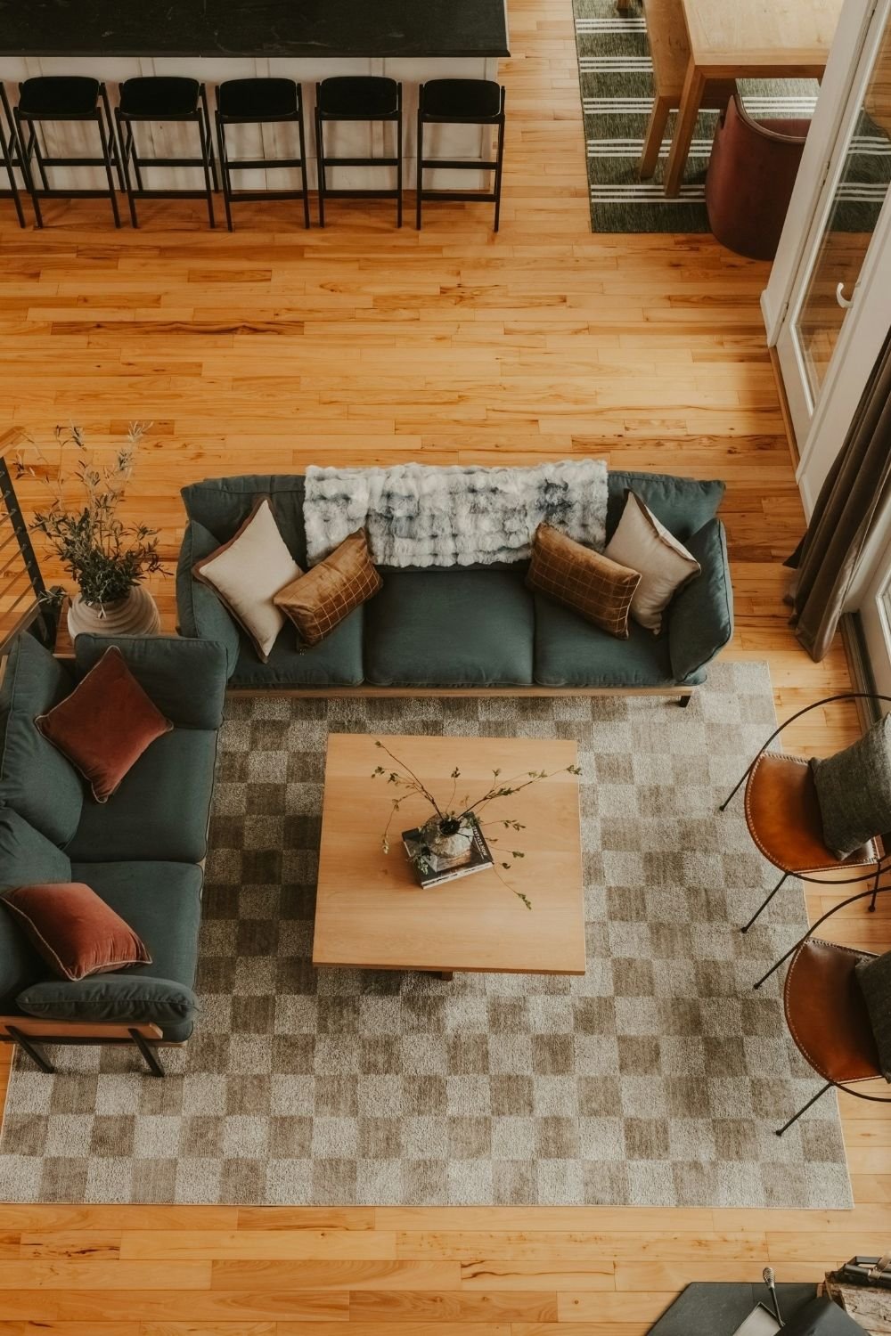 Overhead view of two green sofas in an L with leather chairs, square wood coffee table and checkered rug in an open-plan room