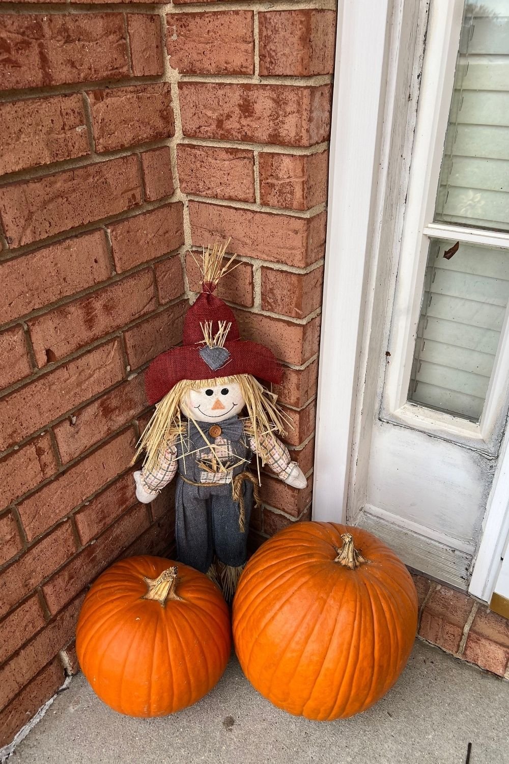 Two orange pumpkins and a fabric scarecrow in the corner of a brick front porch