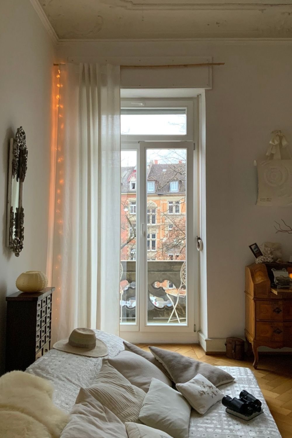 Soft white bedroom with a tall balcony window, linen curtain, fairy lights, layered pillows and an antique wooden desk