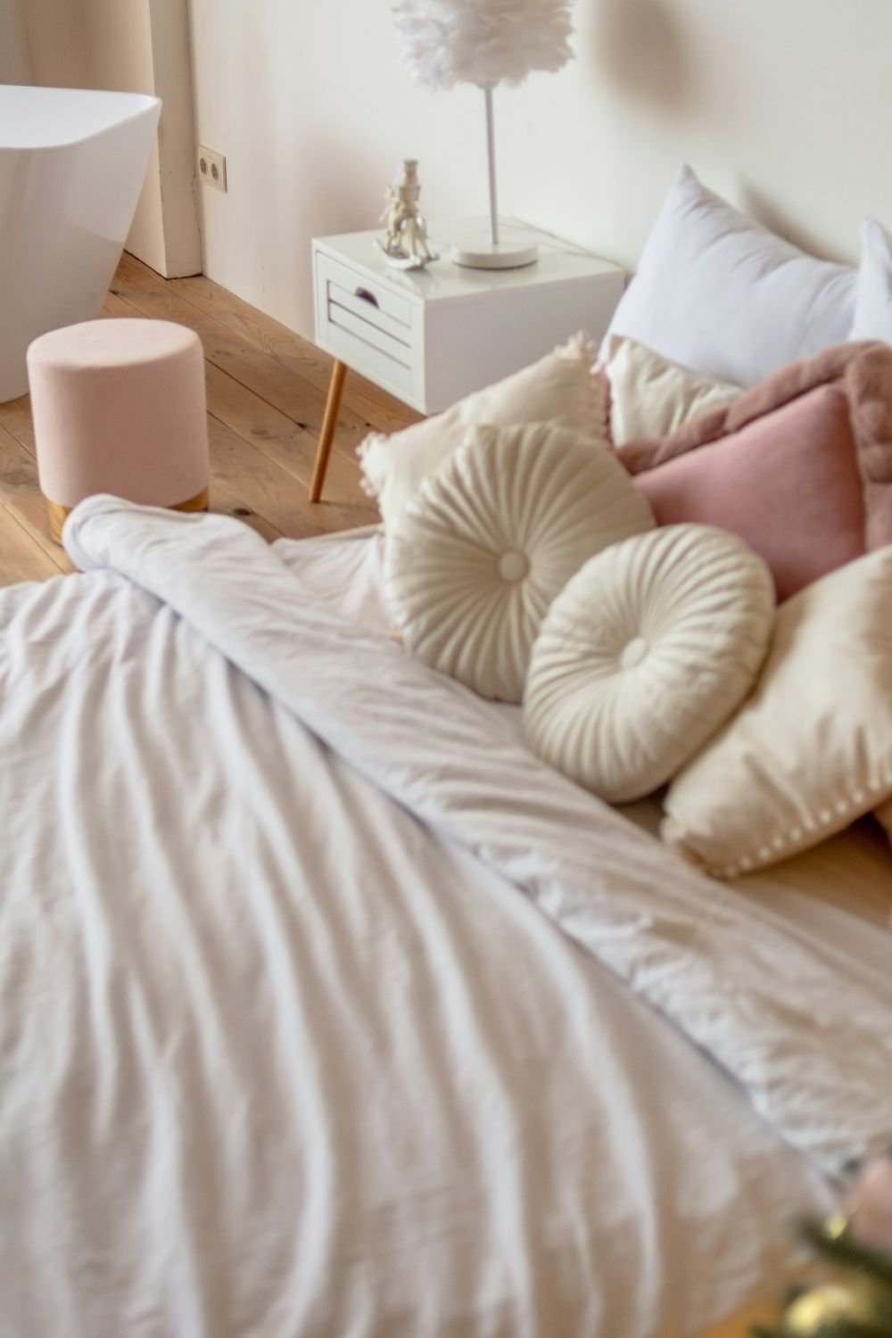 Pastel bedroom with a white duvet, round cream cushions, blush pillows, a pink velvet pouf and a feather lamp