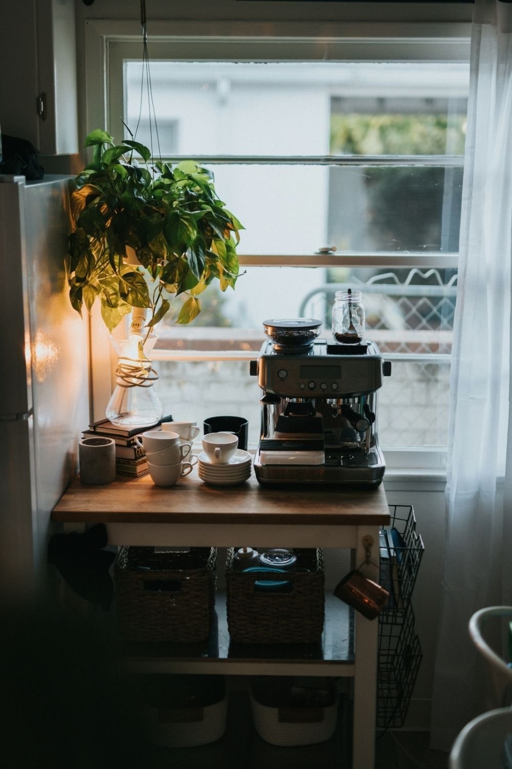 Coffee cart by a kitchen window with an espresso machine, white cups, a pour-over carafe, woven baskets and a hanging pothos