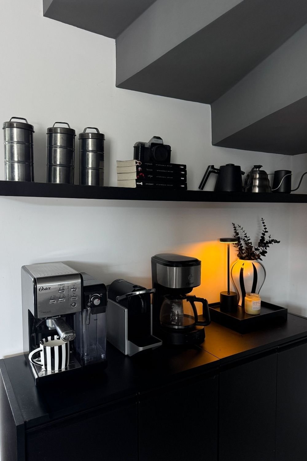 Black coffee station under a staircase with three coffee machines, a shelf of canisters and kettles, and a cordless lamp