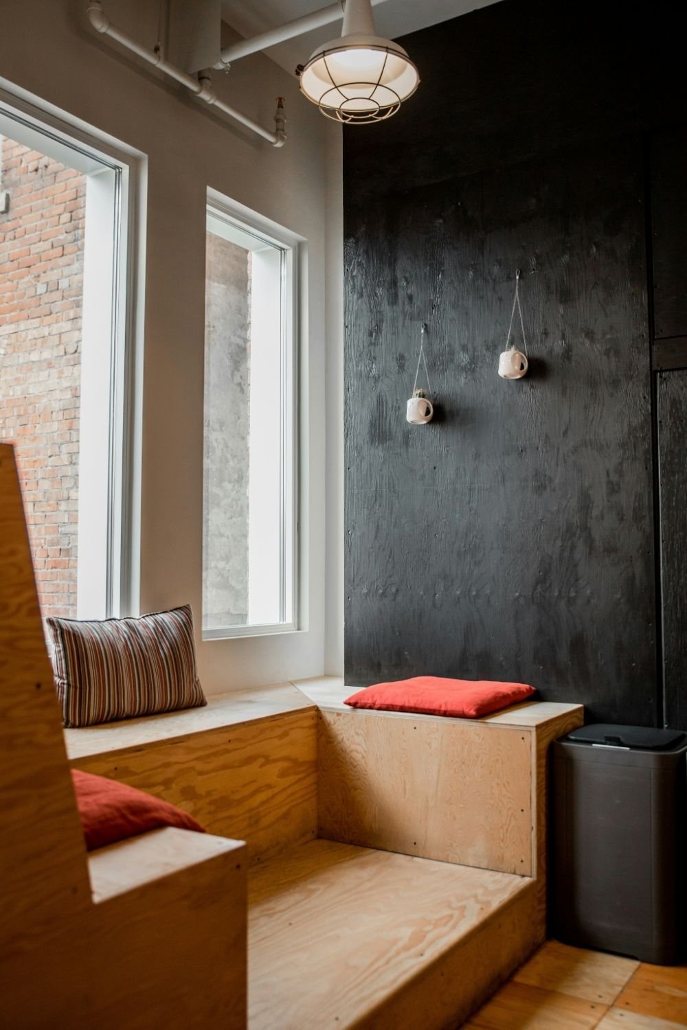 Built-in plywood window bench with striped and red cushions, a black wood wall with hanging planters and a cage pendant light