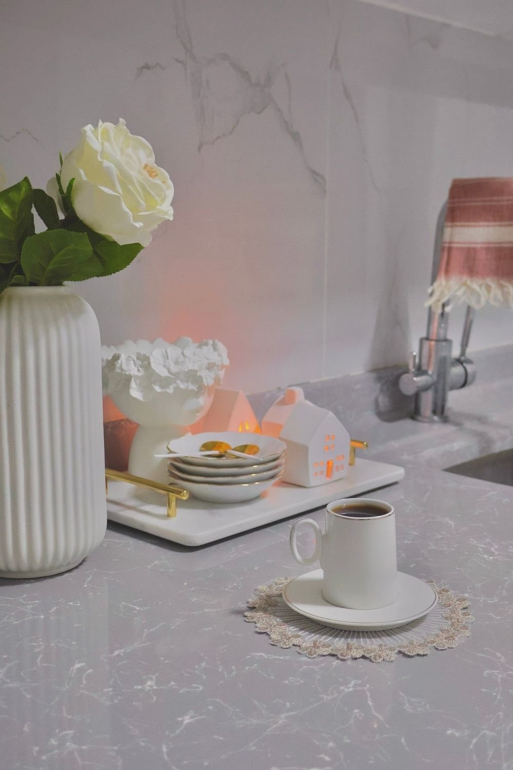 Coffee corner on a gray kitchen counter with a marble tray, gold-rimmed plates, a house lantern and a white rose in a ribbed vase