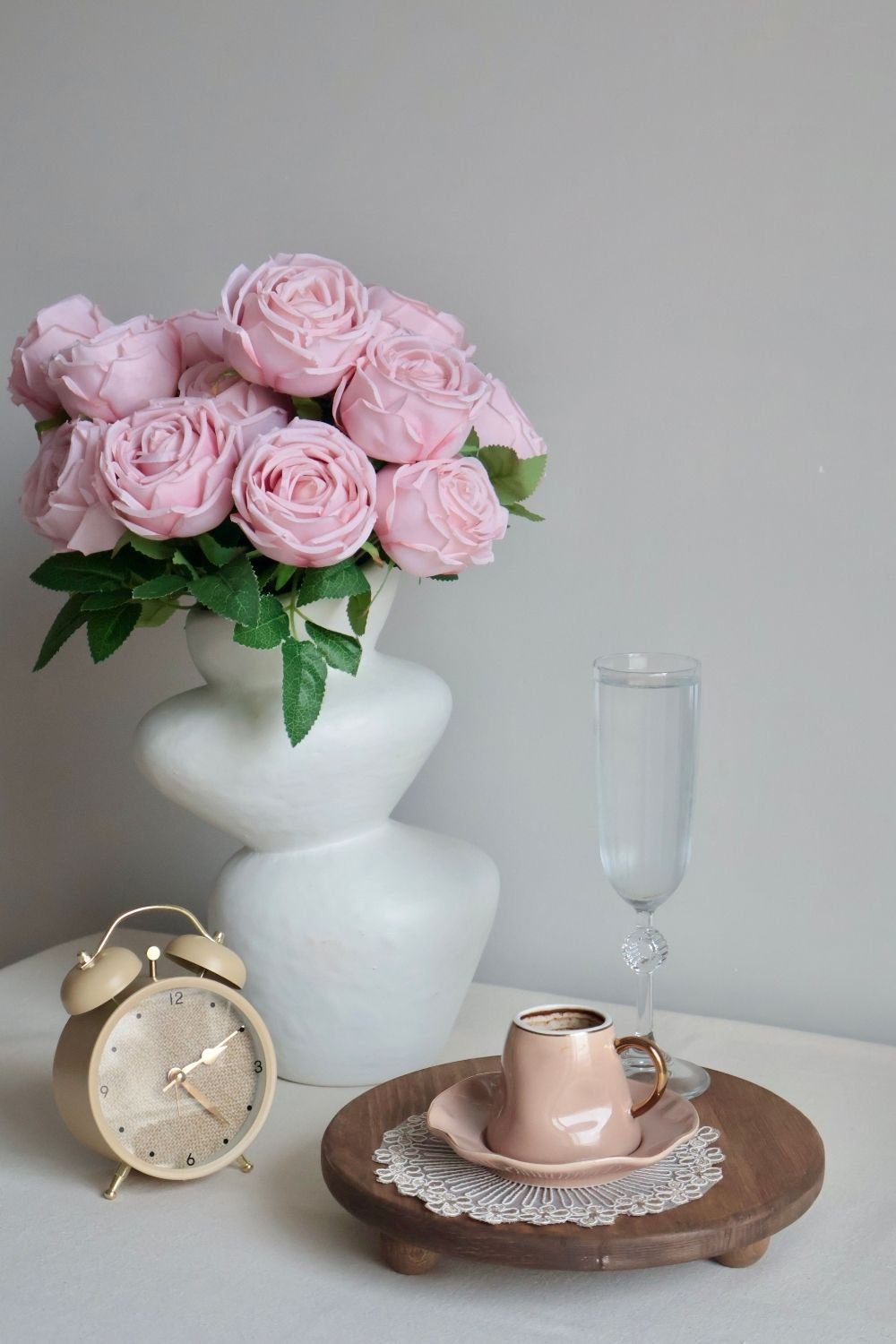 Pink coffee cup on a footed wood riser next to pink roses in a white vase and a small alarm clock