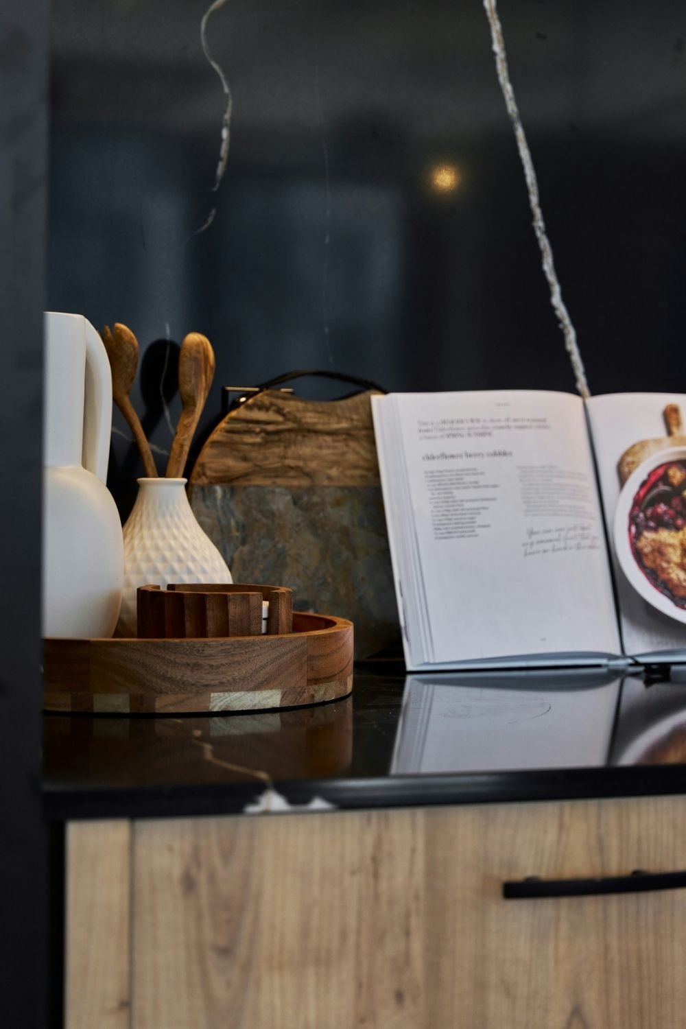 Open cookbook on a dark stone counter next to a round wood tray with a white jug and wooden spoons