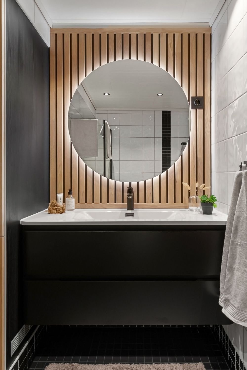 Small bathroom with black paneling, a black floating vanity and oak slats behind a round backlit mirror