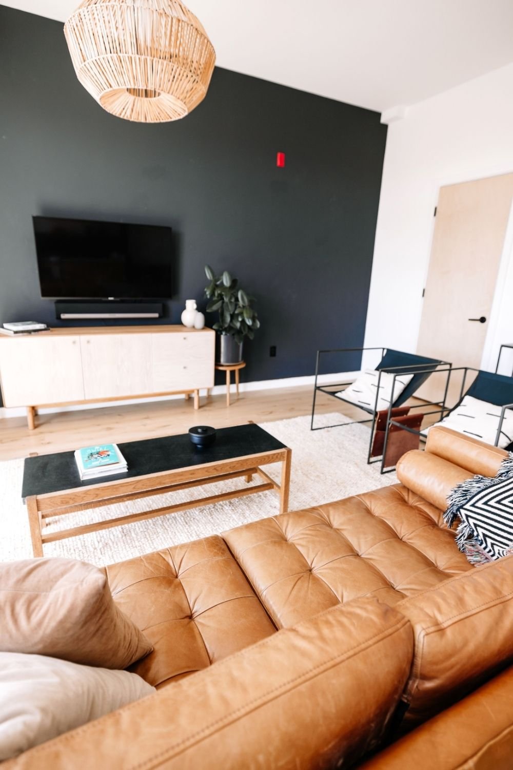 Living room with a black TV wall, pale wood media console, rattan pendant and tan leather sofa