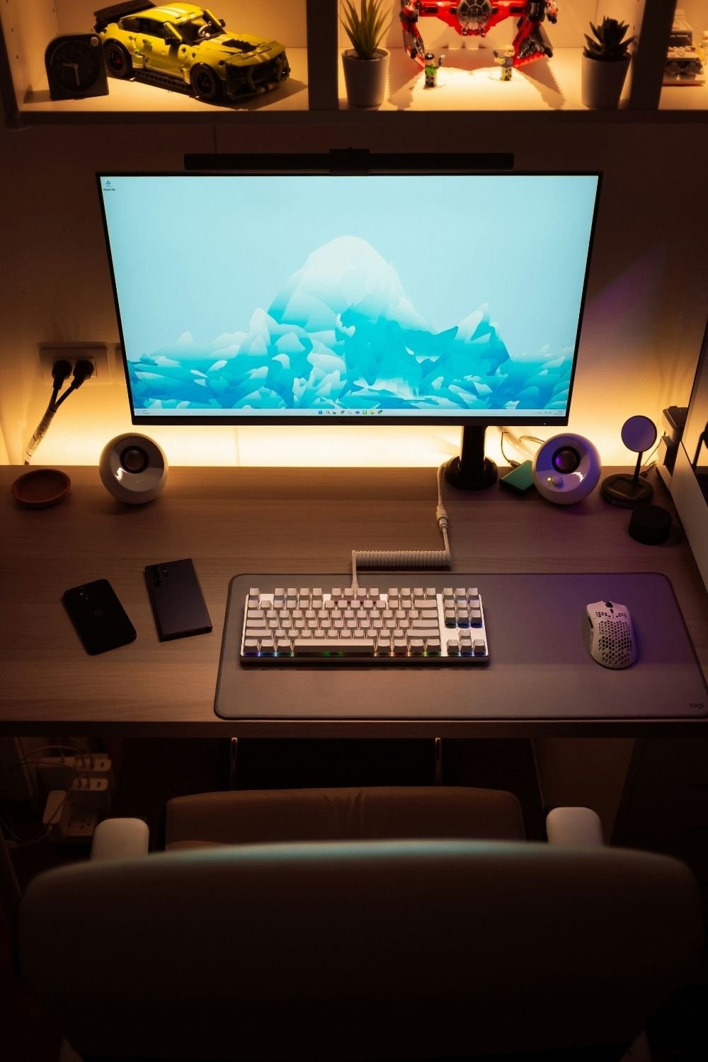 Warm-lit gaming desk setup with an LED strip behind the monitor, white keyboard, gray desk mat and a lit display shelf