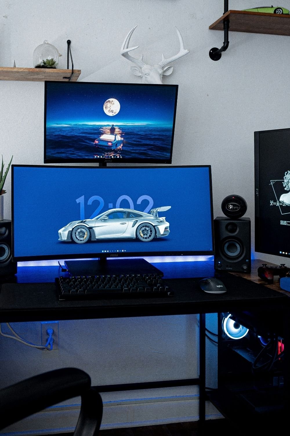 Gaming desk with a curved ultrawide monitor and a second monitor stacked above it, bookshelf speakers and blue LED light