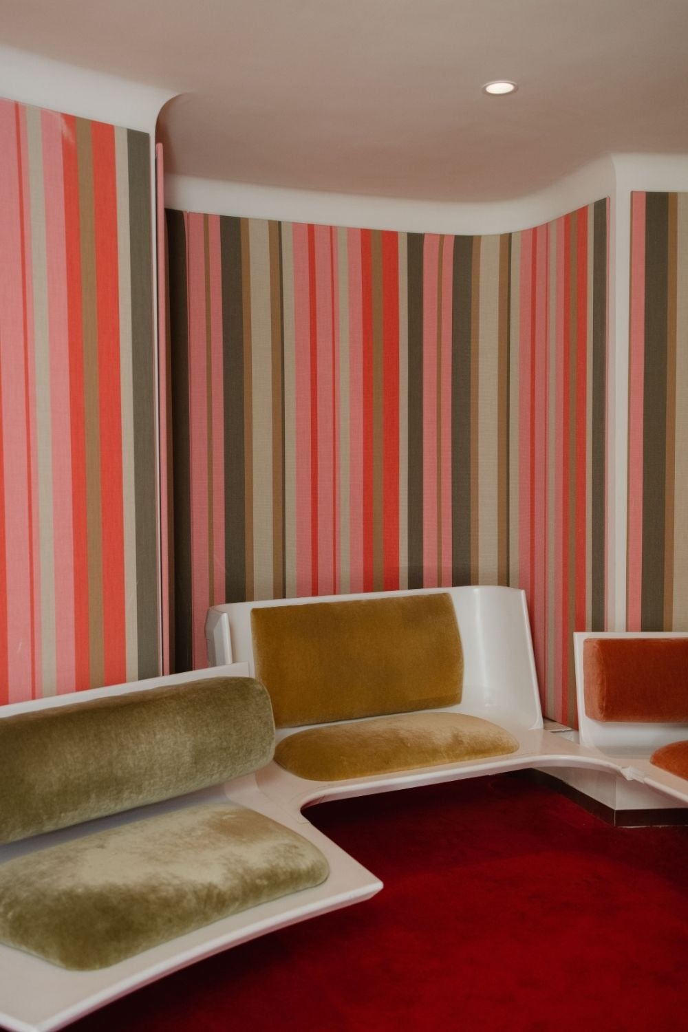 Room with coral, pink, taupe and brown vertical striped walls, molded white benches with velvet cushions and red carpet