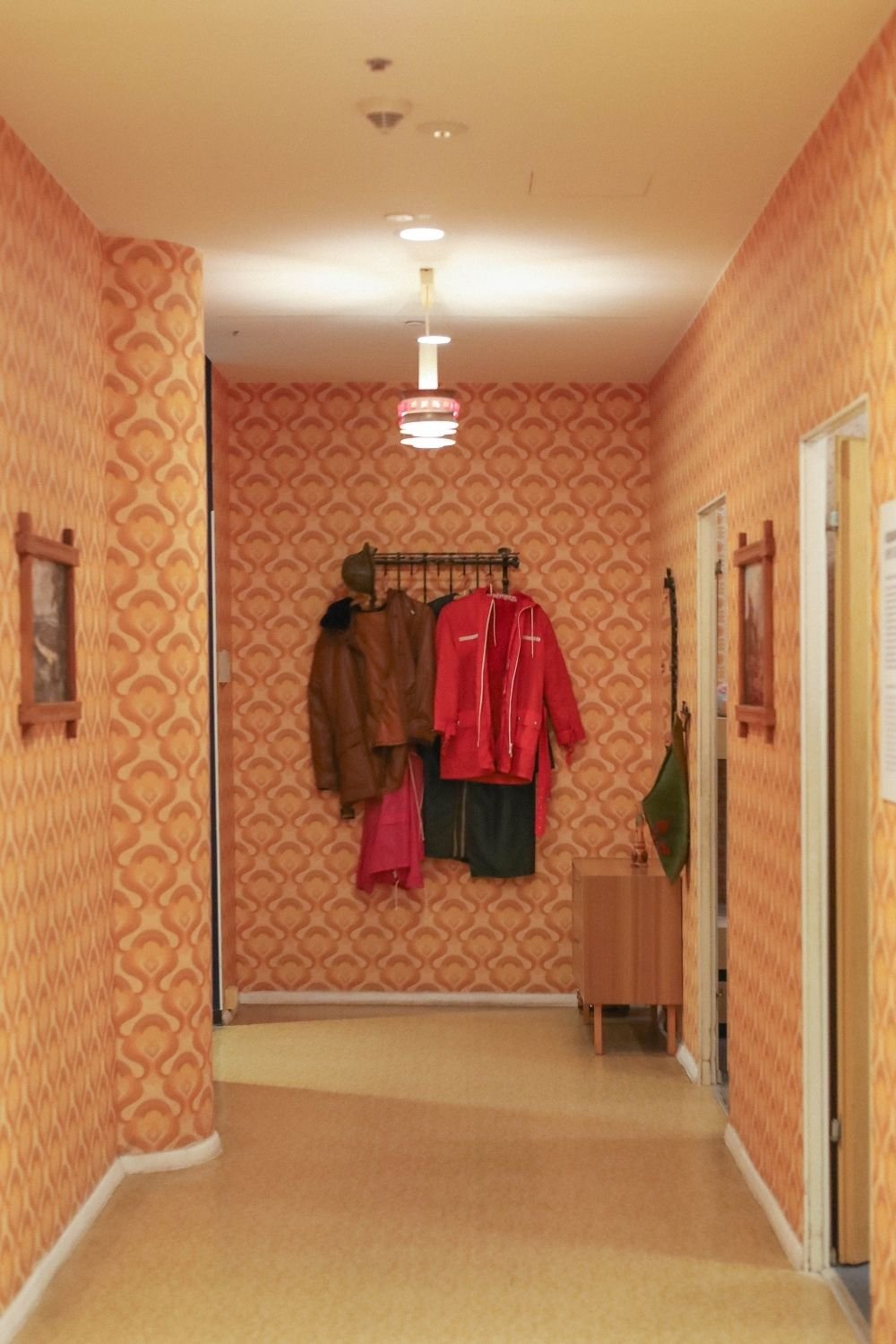 Hallway covered in orange and yellow retro wallpaper with coats on a rail and a vintage ceiling light