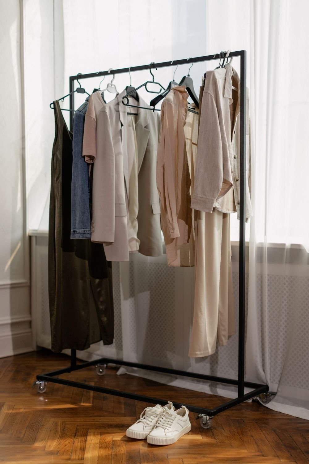 Black freestanding clothing rack on castors by a window, holding neutral shirts, a blazer and trousers