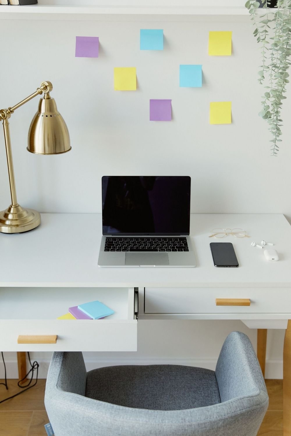 White minimalist desk with a laptop, brass lamp, glasses and sticky notes on the wall above