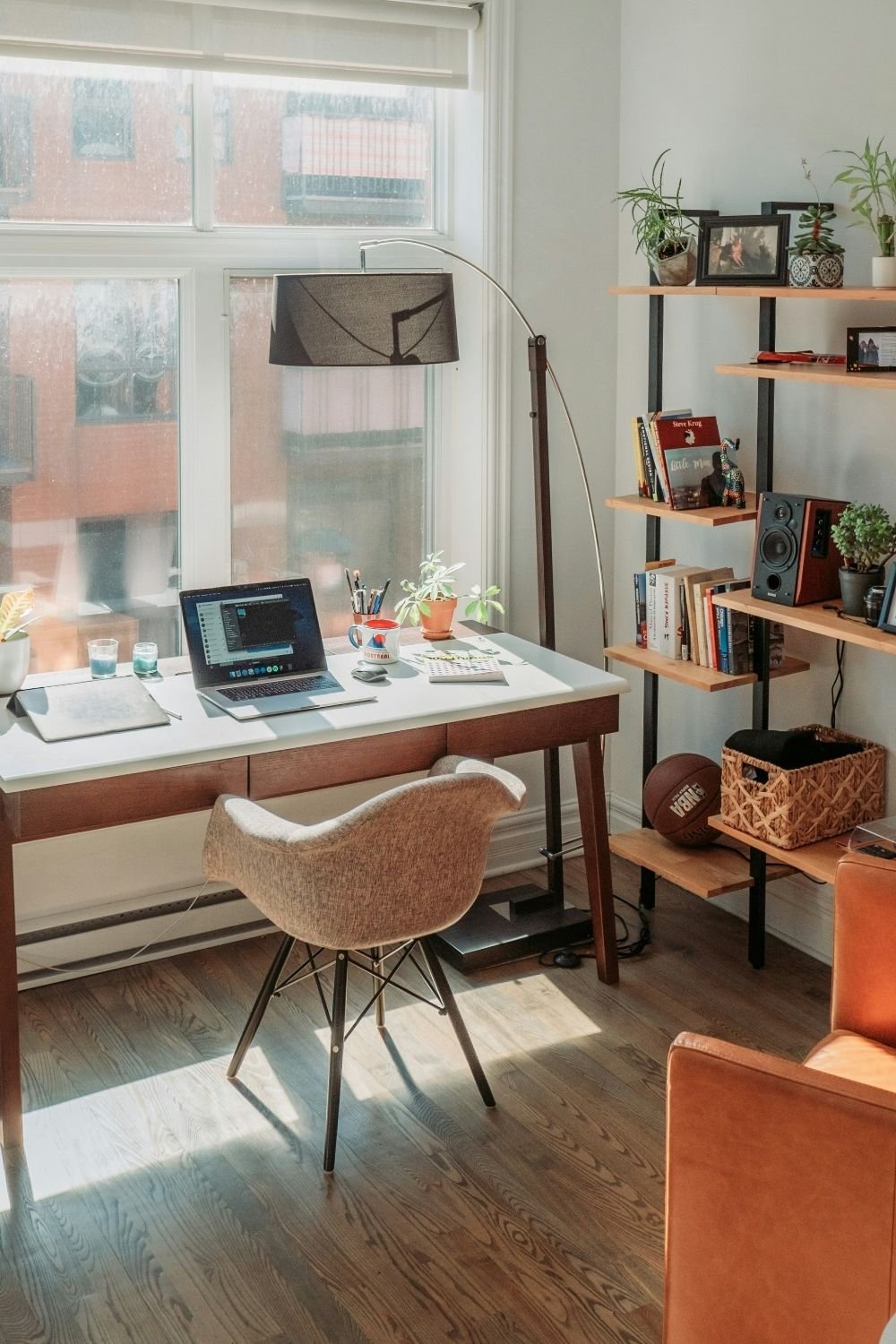 Wood desk under a tall window with an arc floor lamp and an open shelving unit with books and plants