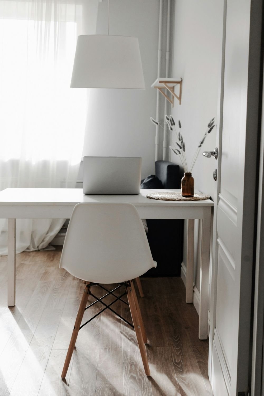 Minimal white desk with a molded chair, laptop and a small vase of dried grasses by a sheer curtain