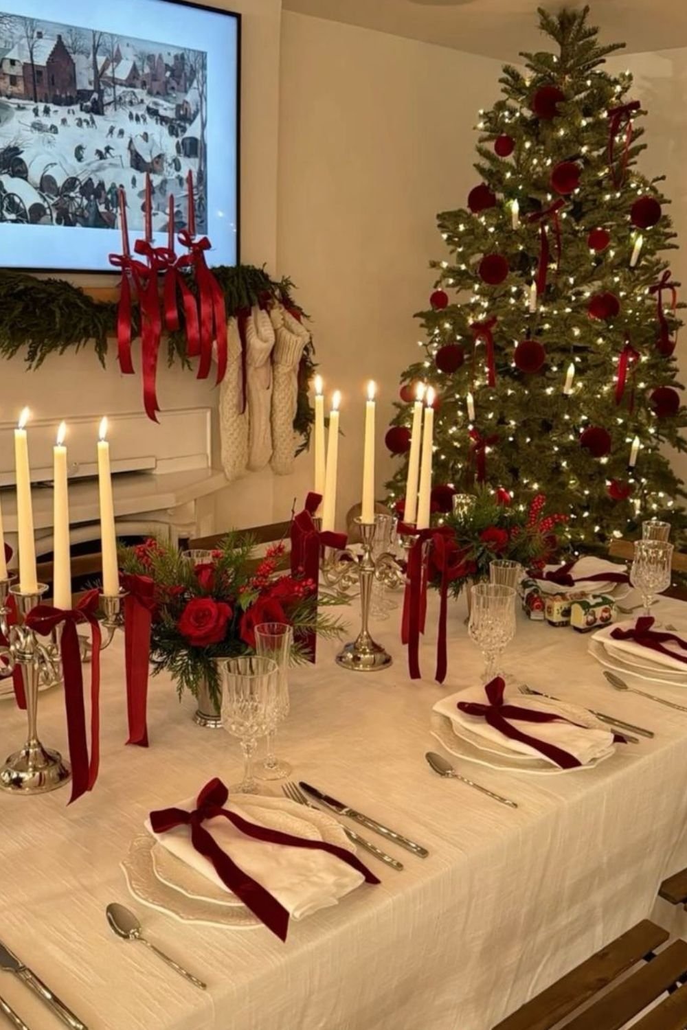 Christmas table with cream linen, red velvet ribbons on napkins, taper candles and red roses, with a decorated tree behind