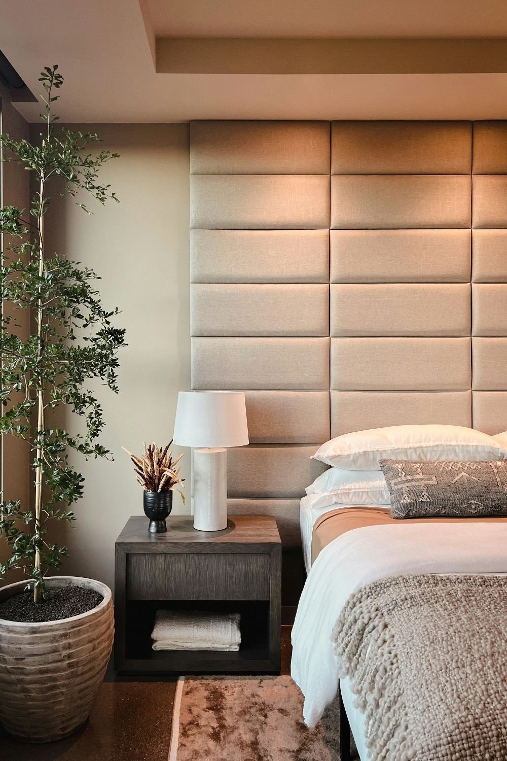 Tall padded sand-colored wall panels behind a bed, with a dark wood nightstand and a potted olive tree