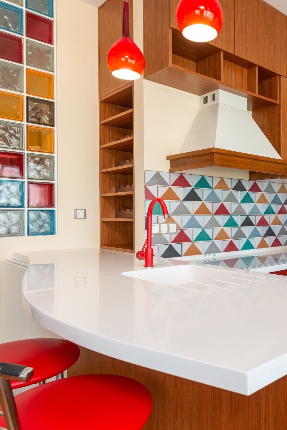 Kitchen with a triangle-patterned backsplash in teal, red and mustard, teak cabinets and red glass pendants