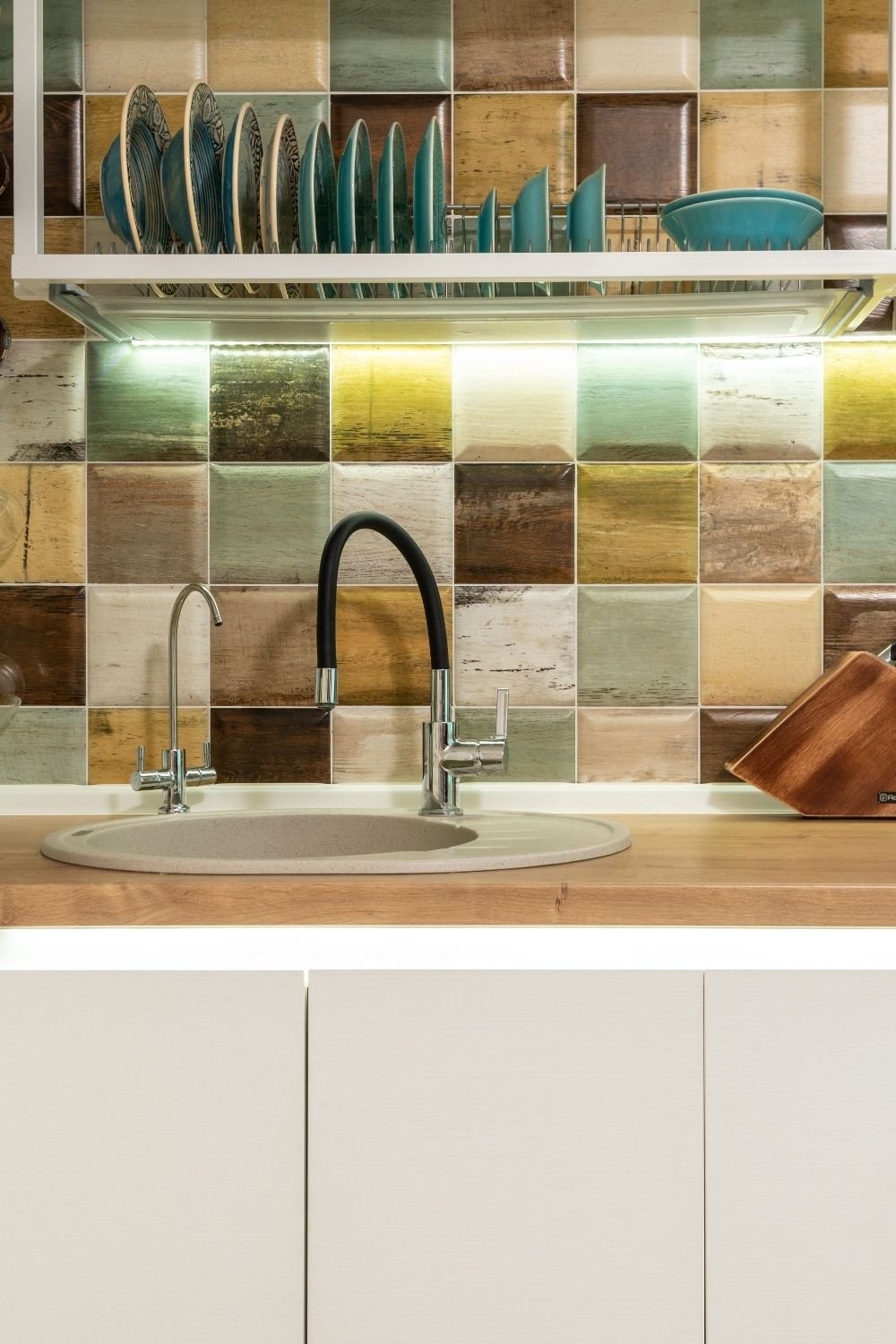 Kitchen backsplash of mixed muted square tiles in green, sand and rust above a wood counter and cream cabinets