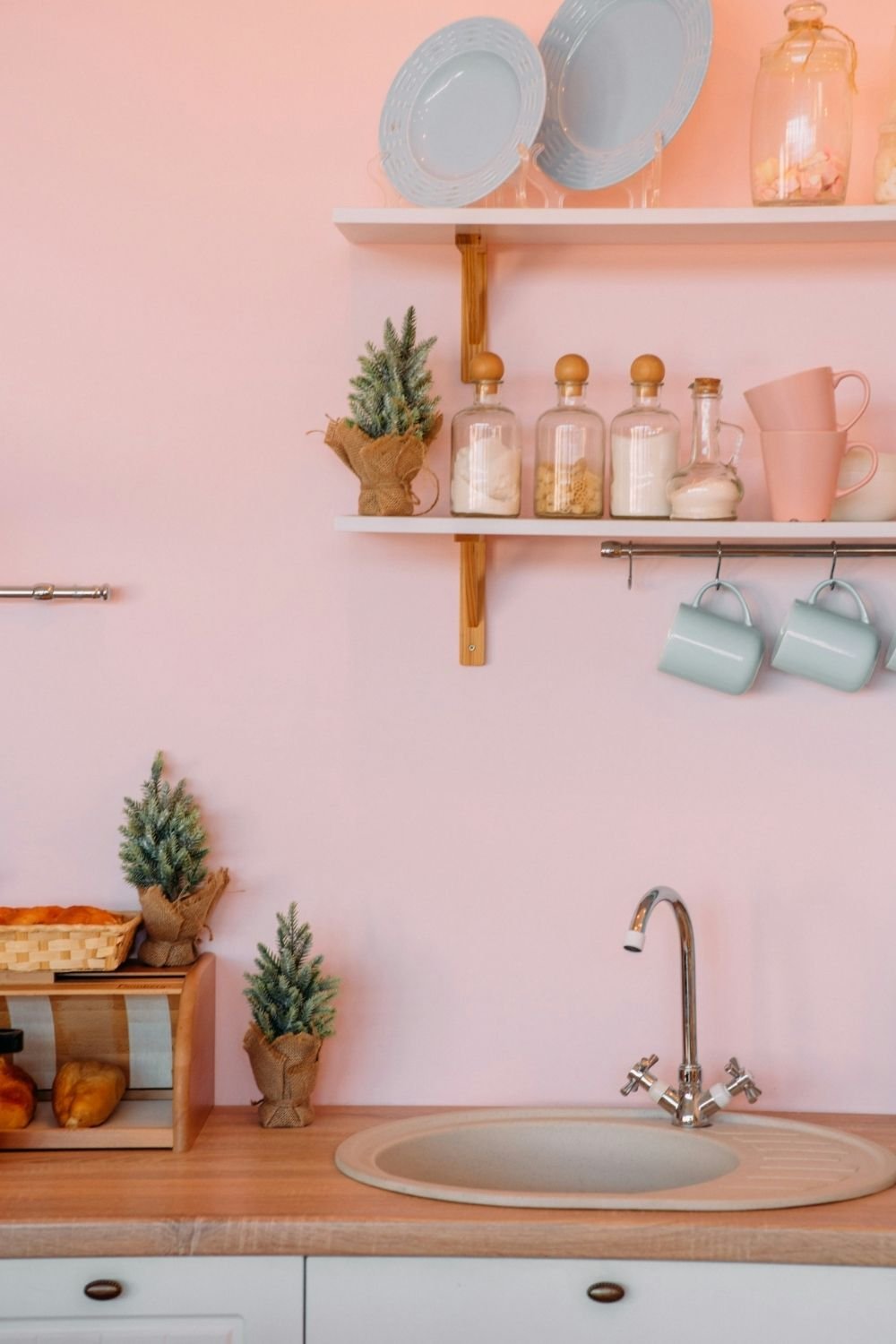 White kitchen with a soft pink wall, pink mugs, sage green cups on a rail and glass jars on open shelves