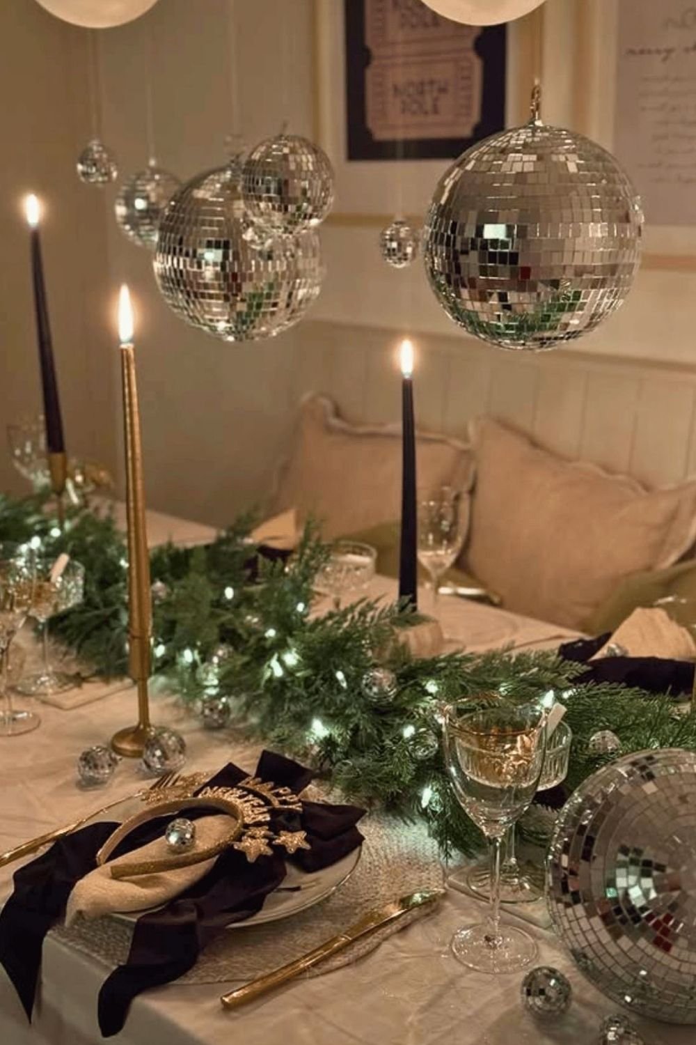 New Year's Eve dinner table with a lit pine garland, black and gold taper candles and disco balls hanging overhead