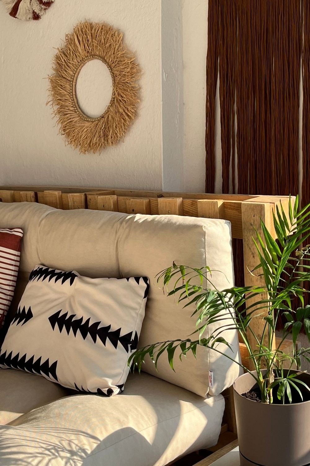 DIY pallet sofa on a balcony with cream and patterned outdoor cushions, a raffia fringed mirror and a palm