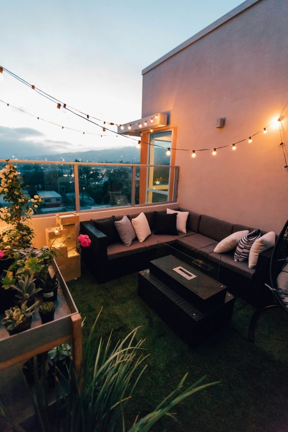 Balcony at dusk lit by two runs of warm festoon bulbs above a dark rattan sectional, turf floor and a low fire table